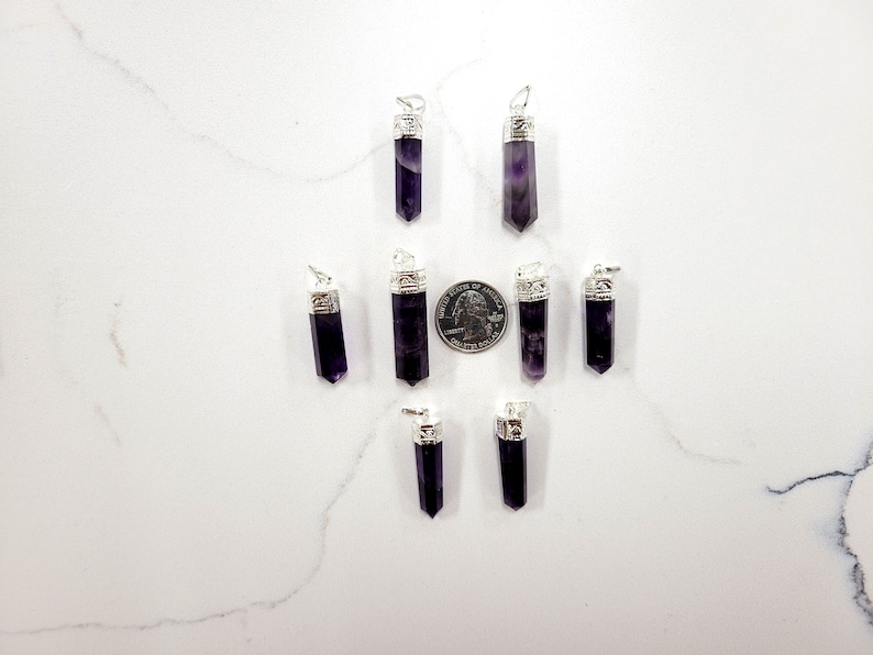 May include: Eight amethyst crystal pendants with silver caps. The pendants are arranged in a star shape on a white surface. A US quarter is included for size reference.