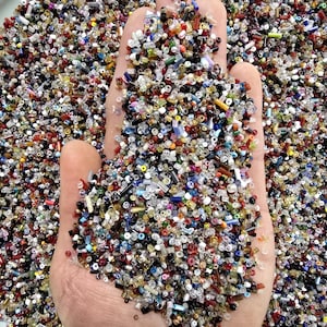 May include: A hand cupped, filled with a multitude of tiny, colorful glass beads. The beads are a mix of colors including red, blue, yellow, and clear, creating a vibrant and textured appearance. The beads are on a white plate.