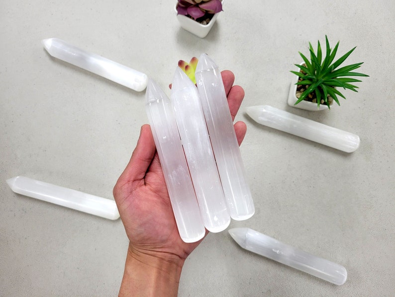 May include: Three white selenite wands with pointed ends, held in a hand. The wands are smooth and polished.