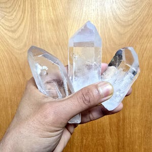 May include: Three clear, hexagonal quartz crystal points with pointed terminations and natural internal inclusions. The crystals are transparent with varying degrees of clarity, featuring smooth, faceted surfaces and distinct geometric shapes.