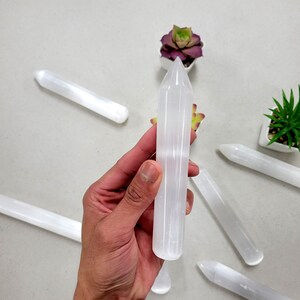May include: A set of six white selenite wands with pointed ends. The wands are smooth and polished, and they are arranged in a circle on a white surface.