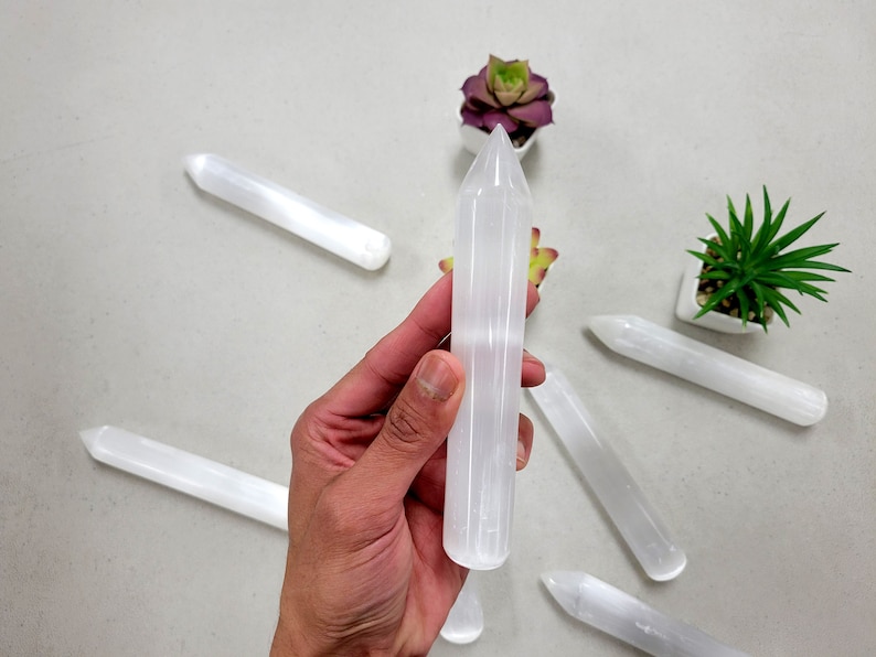 May include: A set of six white selenite wands with pointed ends. The wands are smooth and polished, and they are arranged in a circle on a white surface.