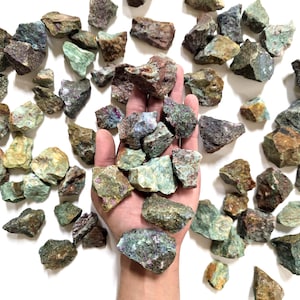May include: A collection of raw, uncut gemstones in shades of green, brown, and purple. The rough stones are held in a hand, with many more scattered on a white surface. The stones vary in size and shape, showcasing natural textures.