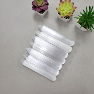 May include: A set of seven white selenite wands, also known as selenite sticks, arranged in a fan shape on a gray surface. The wands are smooth and polished, with a pointed tip and a rounded base.