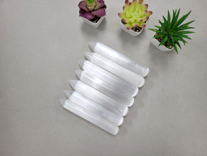 May include: A set of seven white selenite wands, also known as selenite sticks, arranged in a fan shape on a gray surface. The wands are smooth and polished, with a pointed tip and a rounded base.