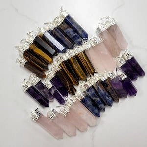 May include: A collection of gemstone pendants in various colors, including amethyst, rose quartz, tiger's eye, and lapis lazuli. Each pendant features a silver-toned cap with an ornate design.