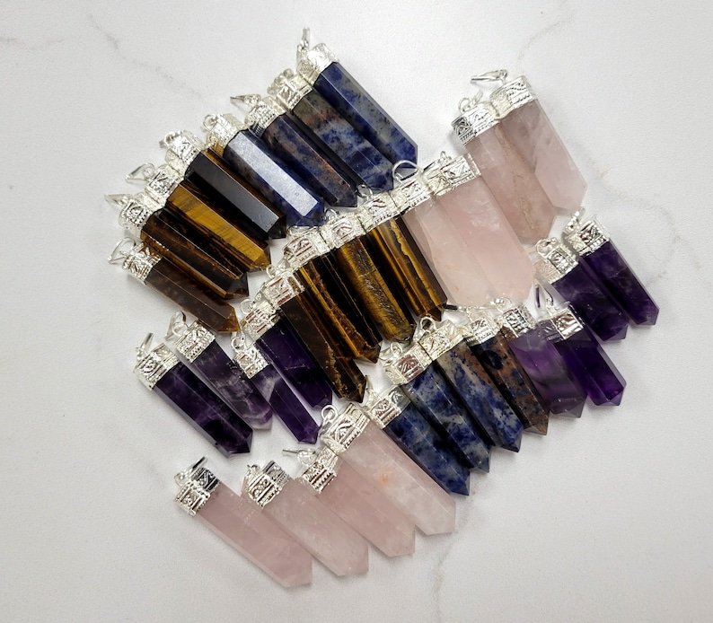 May include: A collection of gemstone pendants in various colors, including amethyst, rose quartz, tiger's eye, and lapis lazuli. Each pendant features a silver-toned cap with an ornate design.