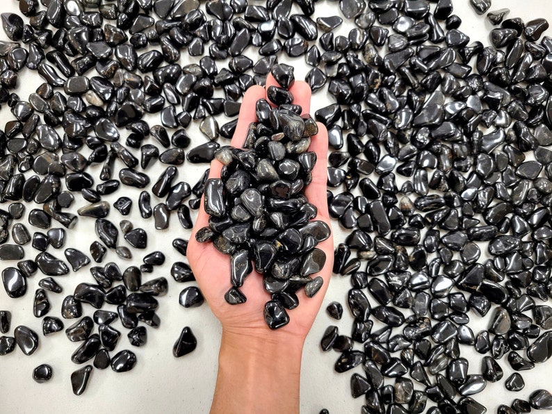 Puede incluir: Una mano sosteniendo un mont&oacute;n de piedras de obsidiana negra pulidas. Las piedras son lisas y brillantes, y est&aacute;n rodeadas de m&aacute;s piedras sobre una superficie blanca.