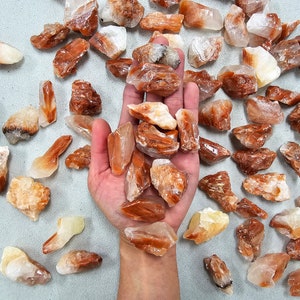 May include: A collection of orange and white calcite crystals. The crystals are rough and have a natural, unpolished look. They are arranged on a white surface.