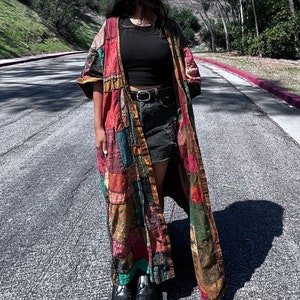 May include: A long, patchwork kimono in shades of red, brown, and green. The kimono is worn over a black top, denim shorts, and black platform boots. The background features a road and trees under a bright blue sky.