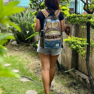 Puede incluir: Una mujer camina por un sendero en un jardín llevando una mochila de mezclilla azul y rayas multicolores.