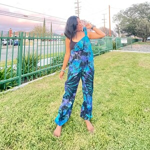 May include: A person wearing a blue and black tie-dye jumpsuit with a black tank top underneath. The person is standing in a grassy area with a green metal fence in the background.