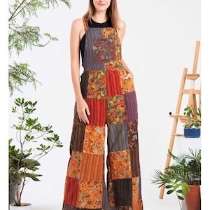 May include: A patchwork jumpsuit featuring a variety of colorful fabric squares. The jumpsuit has wide legs, a bib-style top, and adjustable straps. The patchwork includes floral and striped patterns in shades of orange, red, brown, and purple. The model is wearing a black top underneath.
