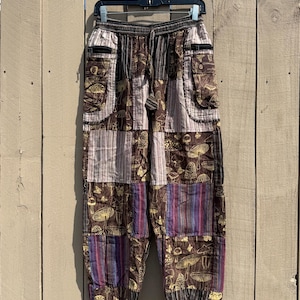 Puede incluir: Pantalones patchwork con estampado de setas marrón y beige, con bolsillos y cintura con cordón. Los pantalones tienen una mezcla de paneles a rayas y estampados en tonos marrón, morado y beige. Cuelgan de una percha negra contra una valla de madera.