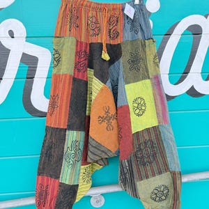 Op de afbeelding: Patchworkbroek met een verscheidenheid aan kleuren, waaronder oranje, rood, groen en zwart. De broek heeft een taille met trekkoord en is versierd met bloemen en geometrische patronen. De broek hangt aan een doorzichtige plastic hanger.