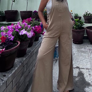 Può includere: Tuta color beige con gambe larghe e parte superiore a pettorina. La tuta presenta una tasca frontale sulla pettorina e chiusure con bottoni sui lati. La modella indossa una canotta bianca sotto. La tuta è realizzata in un tessuto morbido e leggero.