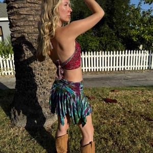 May include: A red paisley halter top and a tie-dye mini skirt with a fringed hem. The outfit is paired with brown knee-high boots. The model is standing outside on a sunny day.