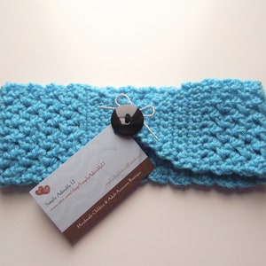 May include: A turquoise blue crocheted headband with a black button and a white ribbon. The headband is handmade and has a tag that says "Simply Adorable II" and "Handmade Children & Adult Accessory Boutique".
