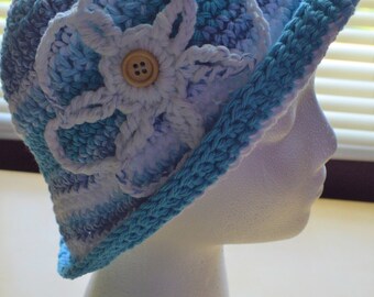 May include: A blue and white crocheted hat with a flower design and a brown button. The hat is on a white mannequin head.