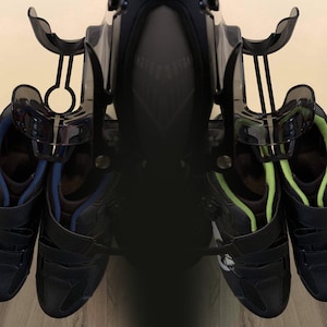 May include: Two pairs of black cycling shoes with velcro straps, one pair with blue accents and the other with green accents, hanging on a black shoe rack.