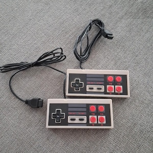 May include: Two retro-style video game controllers with black directional pads, gray select and start buttons, and red action buttons. The controllers are off-white with black accents and connected by black cords.