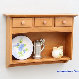 May include: A wooden wall shelf with three drawers and a lower shelf. The shelf is painted a light brown color and has a decorative curved top. The lower shelf has a plate with blue flowers, a silver pitcher, and a white pitcher with pink flowers.