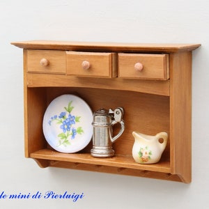 May include: A wooden wall shelf with three drawers and a lower open shelf. The shelf is decorated with a white plate with blue flowers, a silver pitcher, and a white pitcher with pink flowers.