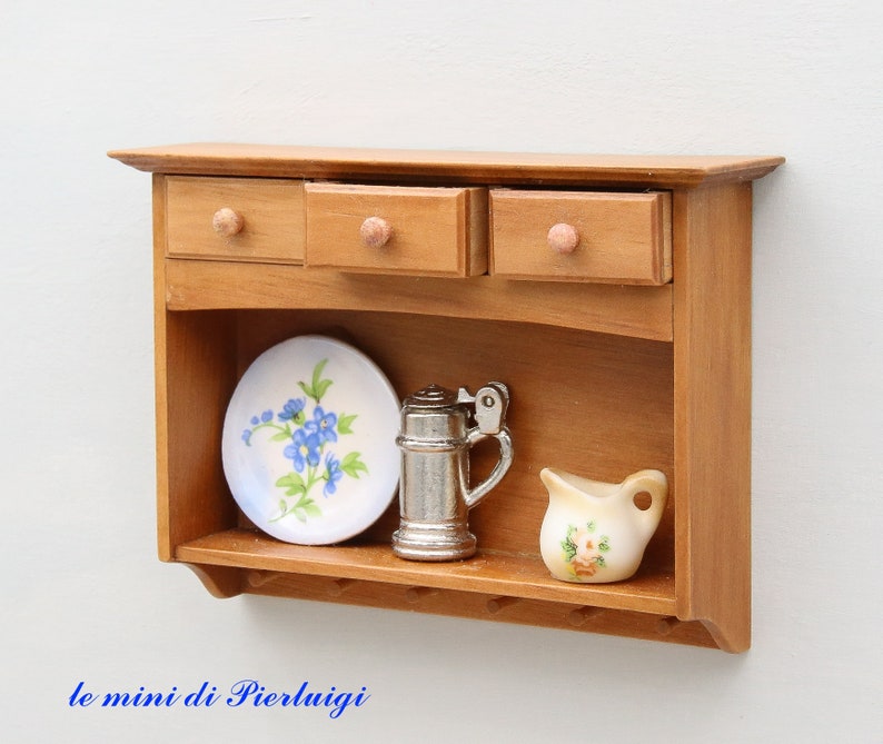 May include: A wooden wall shelf with three drawers and a lower open shelf. The shelf is decorated with a white plate with blue flowers, a silver pitcher, and a white pitcher with pink flowers.