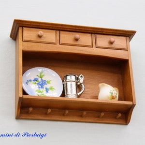 May include: A wooden wall shelf with three drawers and a lower shelf. The shelf has hooks on the bottom and is decorated with a blue and white plate, a silver pitcher, and a small white pitcher with floral designs.