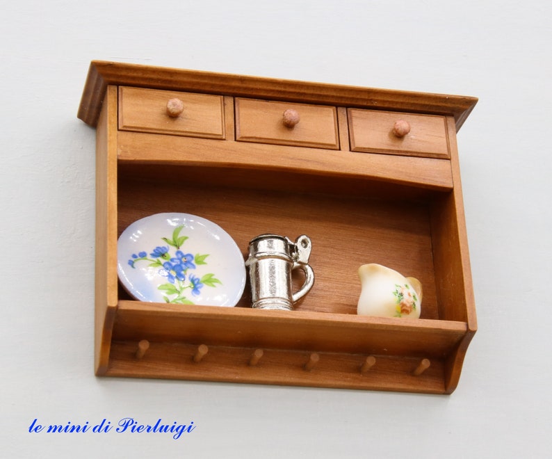 May include: A wooden wall shelf with three drawers and a lower shelf. The shelf has hooks on the bottom and is decorated with a blue and white plate, a silver pitcher, and a small white pitcher with floral designs.