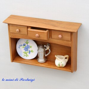 May include: A miniature wooden cabinet with three drawers and a shelf. The shelf has a blue and white floral plate, a silver pitcher, and a small white pitcher with floral designs.