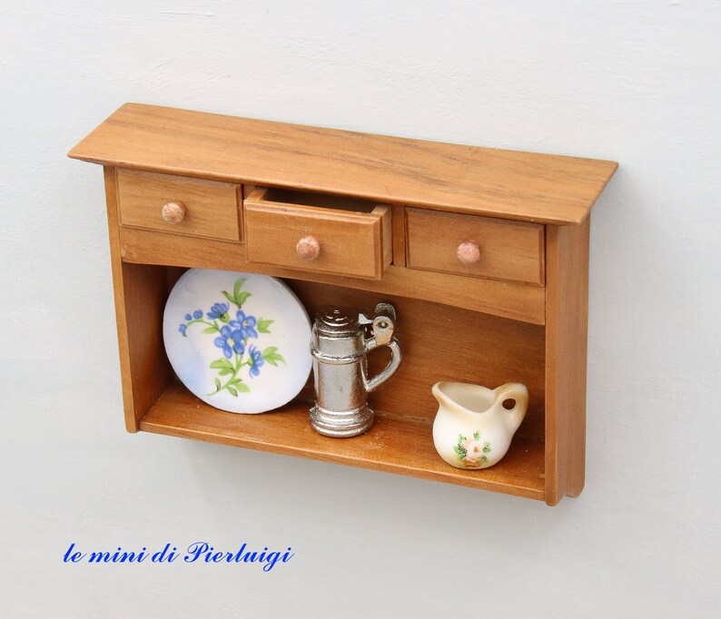 May include: A miniature wooden cabinet with three drawers and a shelf. The shelf has a blue and white floral plate, a silver pitcher, and a small white pitcher with floral designs.