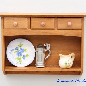 May include: A wooden wall shelf with three drawers and a lower shelf. The shelf has a floral patterned plate, a silver pitcher, and a small white pitcher with floral designs.
