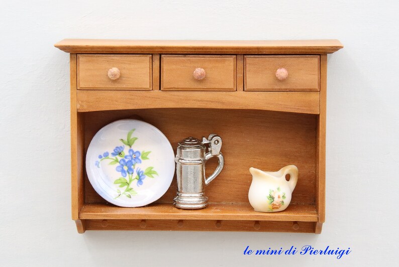 May include: A wooden wall shelf with three drawers and a lower shelf. The shelf has a floral patterned plate, a silver pitcher, and a small white pitcher with floral designs.
