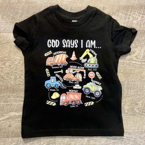 Peut inclure: T-shirt noir avec un motif graphique de véhicules de chantier et le texte "God Says I Am..." avec des versets bibliques.