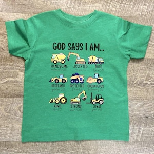 Peut inclure: T-shirt vert avec un graphique de véhicules de chantier et le texte "GOD SAYS I AM..." avec des versets bibliques et des mots comme "handsome", "accepted", "bold", "redeemed", "protected", "courageous", "kind", "strong", et "loyal".