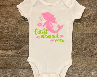 Peut inclure: Combinaison blanche pour bébé avec une sirène en paillettes roses et un texte en paillettes vertes qui dit "Cutest mermaid ever".