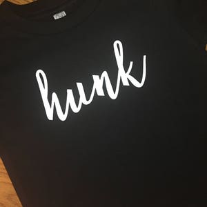 May include: Black t-shirt with the white word "hunk" in a cursive font. The shirt has a crew neck. The image is a close-up shot of the shirt. The word "hunk" is written in a stylish, flowing script.