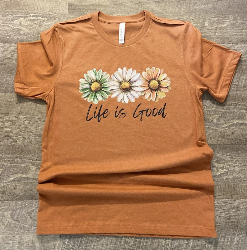 May include: An orange t-shirt featuring three daisy flowers in green, white, and yellow, with the phrase "Life is Good" printed below. The shirt has a crew neck and short sleeves, and is made of a soft material.