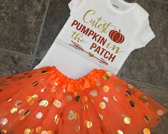 May include: A white baby bodysuit with the text "Cutest Pumpkin in the Patch" in gold glitter and brown lettering, paired with an orange tutu skirt with gold and silver polka dots, and an orange floral headband.