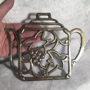 May include: A brass teapot-shaped hot pad with a grapevine design. The pad is intricately detailed with leaves and grapes.