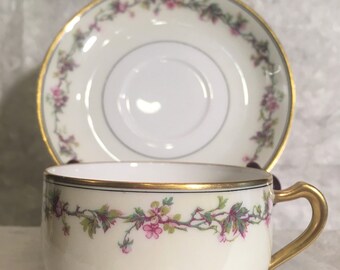 May include: A white teacup and saucer with a gold rim and a floral pattern of pink flowers and green leaves. The pattern is a delicate vine design.