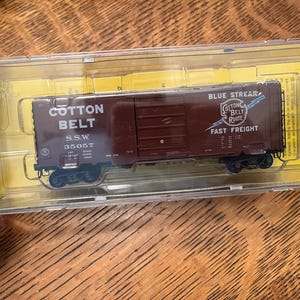 May include: A brown Cotton Belt Route model train car in a clear plastic case. The car has the words "Cotton Belt" and "Blue Streak Fast Freight" printed on the side. The model train car is a replica of a freight car.