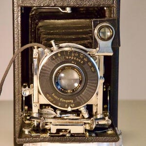May include: A vintage black and silver Speedex No. 1 camera with a lens and a cable release. The camera has a bellows design and is marked with the Ansco Company logo. The camera is open, revealing its internal components.