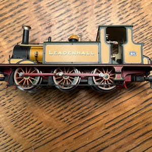 May include: A detailed model train locomotive with the name "LEADENHALL" printed on the side. The train features a black smokestack, gold and green accents, and red wheels. The model sits on a wooden surface.