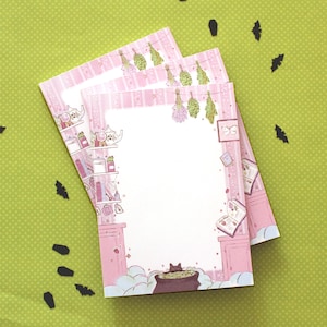 May include: A stack of pink and white notepad paper with a black cat illustration and a floral design. The paper has a pink and white striped border. The paper is decorated with a black cat, a cauldron, and a variety of herbs and flowers.