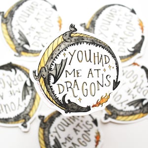 Op de afbeelding: Een sticker met een zwarte en grijze draak die om een cirkel is gekruld met de tekst "You had me at dragons" in gouden letters. De draak blaast vuur en er zijn gouden sterren rond de tekst.