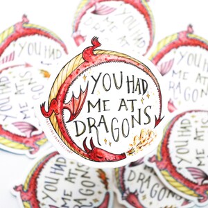 Op de afbeelding: Een rode draak met gele accenten blaast vuur en wikkelt zich om een witte cirkel met de tekst "You had me at dragons".