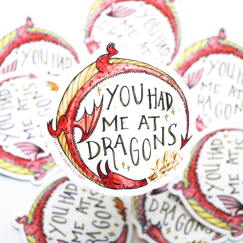 Op de afbeelding: Een rode draak met gele accenten blaast vuur en wikkelt zich om een witte cirkel met de tekst "You had me at dragons".