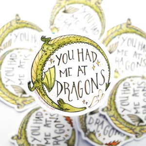 Op de afbeelding: Een ronde sticker met een groene draak die om de tekst "You had me at dragons" gekruld is. De sticker heeft een witte achtergrond met gouden glitteraccenten.